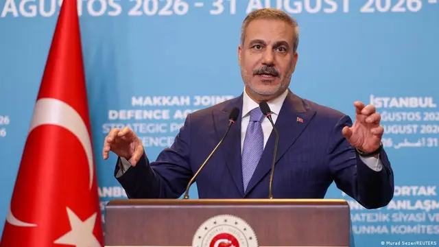 Dışişleri Bakanı Hakan Fidan kürsüde konuşuyor. İki elini havaya kaldırmış, arkasında Türkiye bayrağı ve mavi fon var